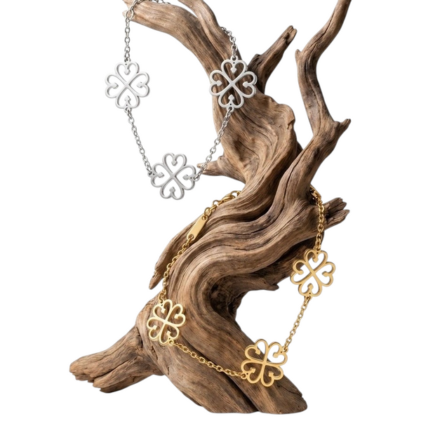 Aureliss Armband Nyame mit filigranen Kleeblatt-Motiven in Gold und Silber auf einem dekorativen Treibholz-Ständer.